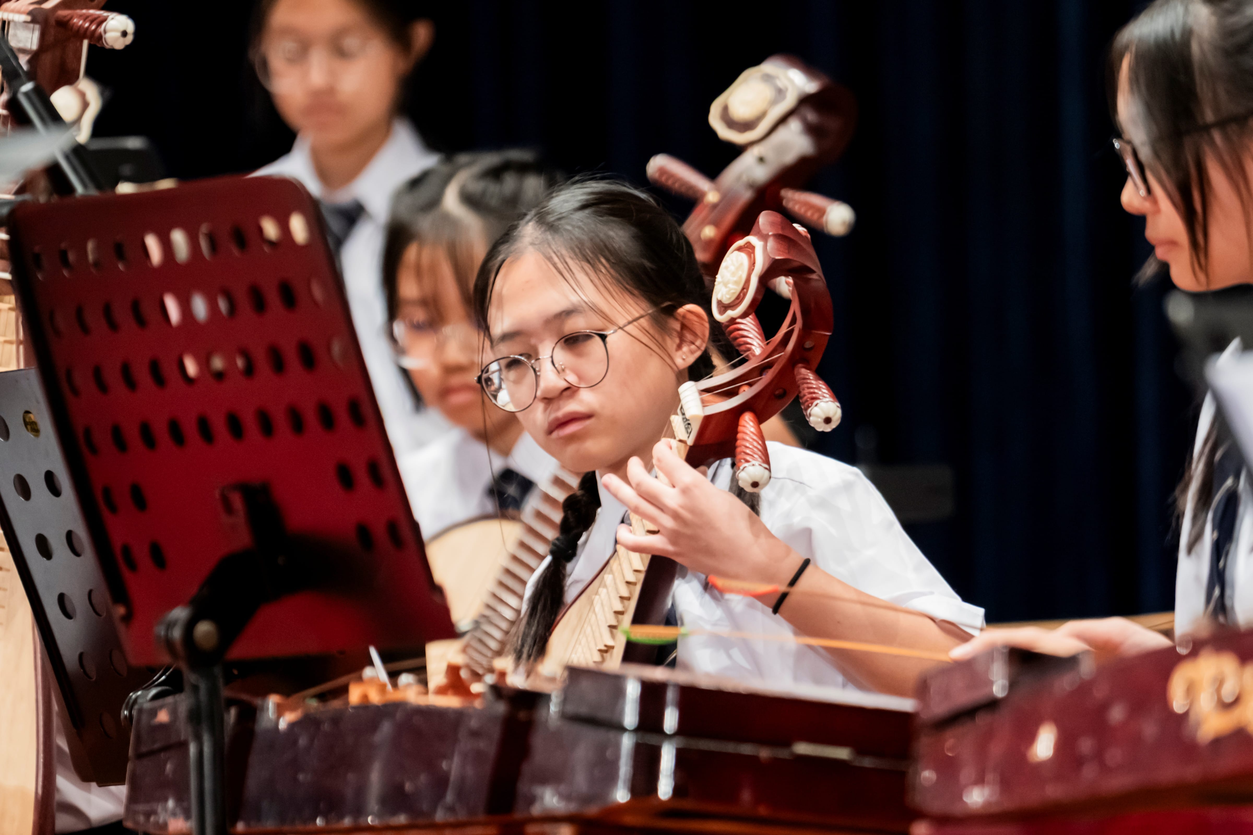 A female student wearing glasses attentively plays a pipa during a Chinese orchestra performance.